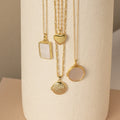 Four gold locket necklaces hang vertically on a cream textured vase—featuring rectangular, heart, and two round lockets, some with smooth polished finishes and others with glossy mother-of-pearl inlays.