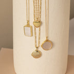 Four gold locket necklaces hang vertically on a cream textured vase—featuring rectangular, heart, and two round lockets, some with smooth polished finishes and others with glossy mother-of-pearl inlays.