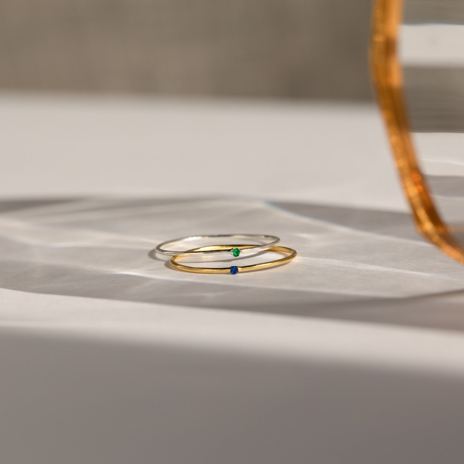 Two Thin Mini Birthstone Rings—one gold with a blue stone, one silver with a green stone—rest on a white surface, ideal for minimalists or as a Christmas gift.