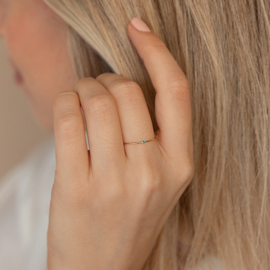 A hand adorned with the Thin Mini Birthstone Ring, its petite blue stone shining, brushes through long blonde hair—making it an ideal minimalist accessory and a thoughtful Christmas gift.