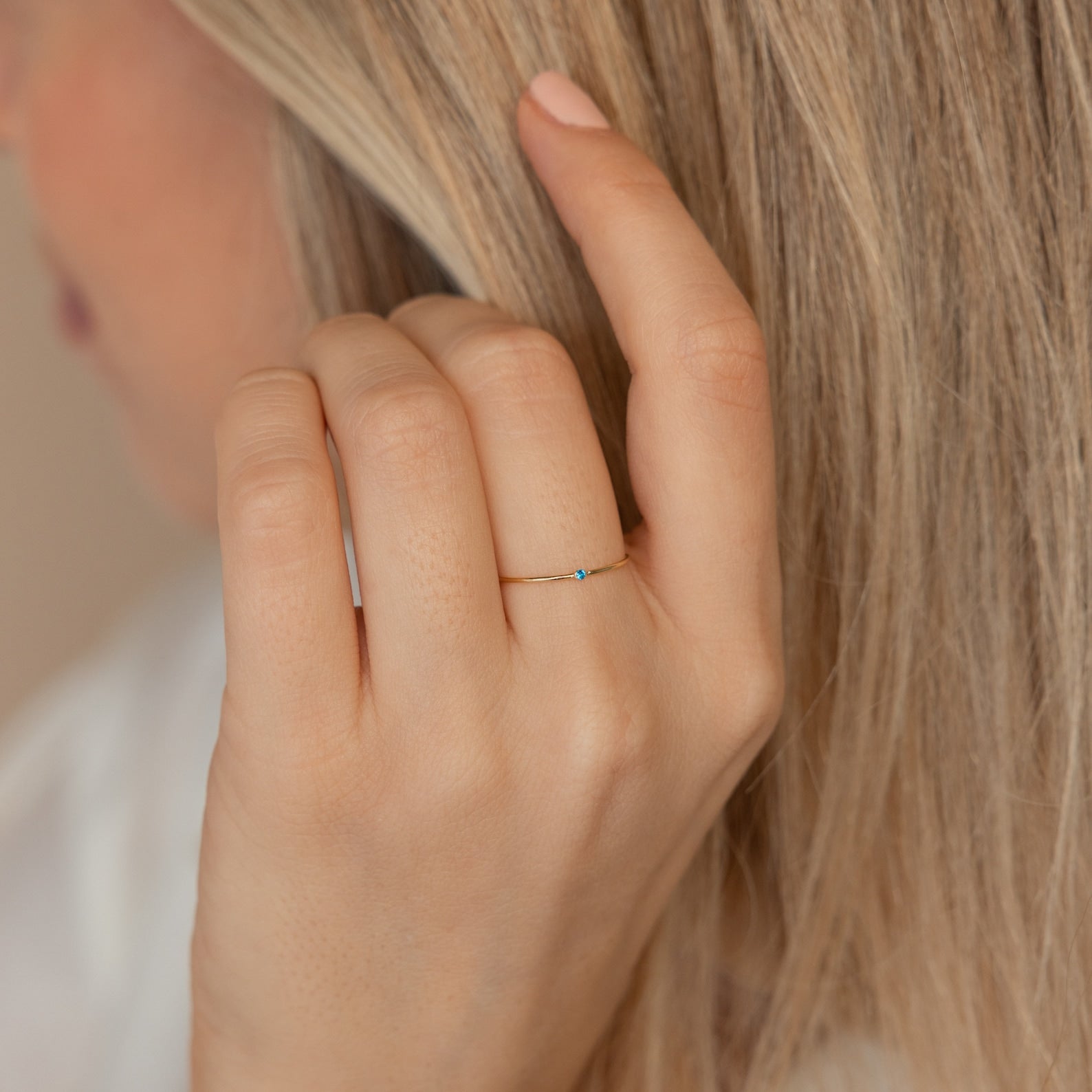 A hand adorned with the Thin Mini Birthstone Ring, its petite blue stone shining, brushes through long blonde hair—making it an ideal minimalist accessory and a thoughtful Christmas gift.