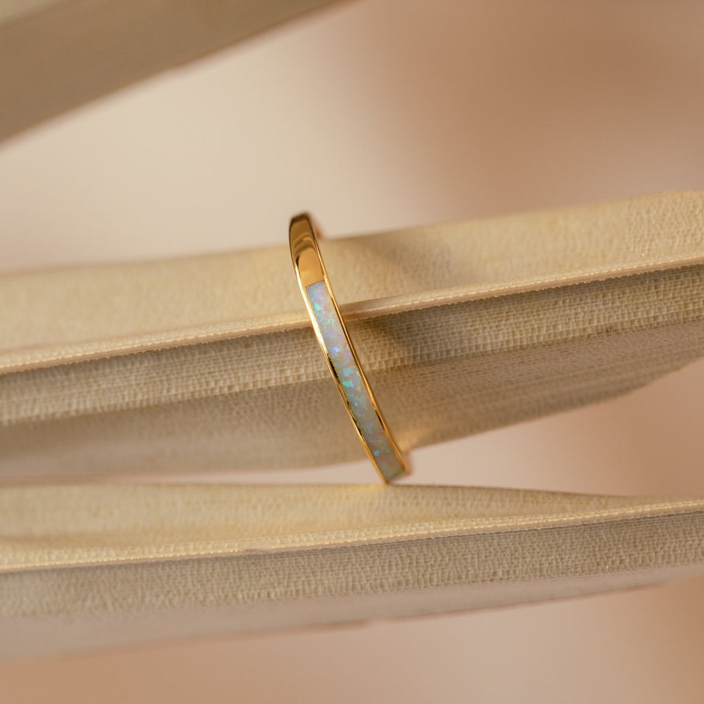 The White Opal Inlay Band in 18K Gold rests on the pages of an open book, showcasing its striking gemstone design.
