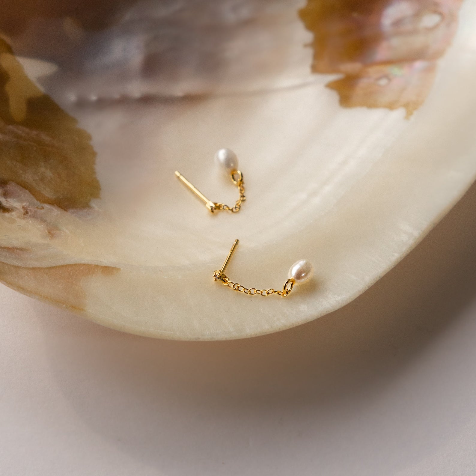 The Isha Pearl Drop Earrings in 18K Gold, with delicate chains and small pearls, are displayed elegantly on a large iridescent seashell.
