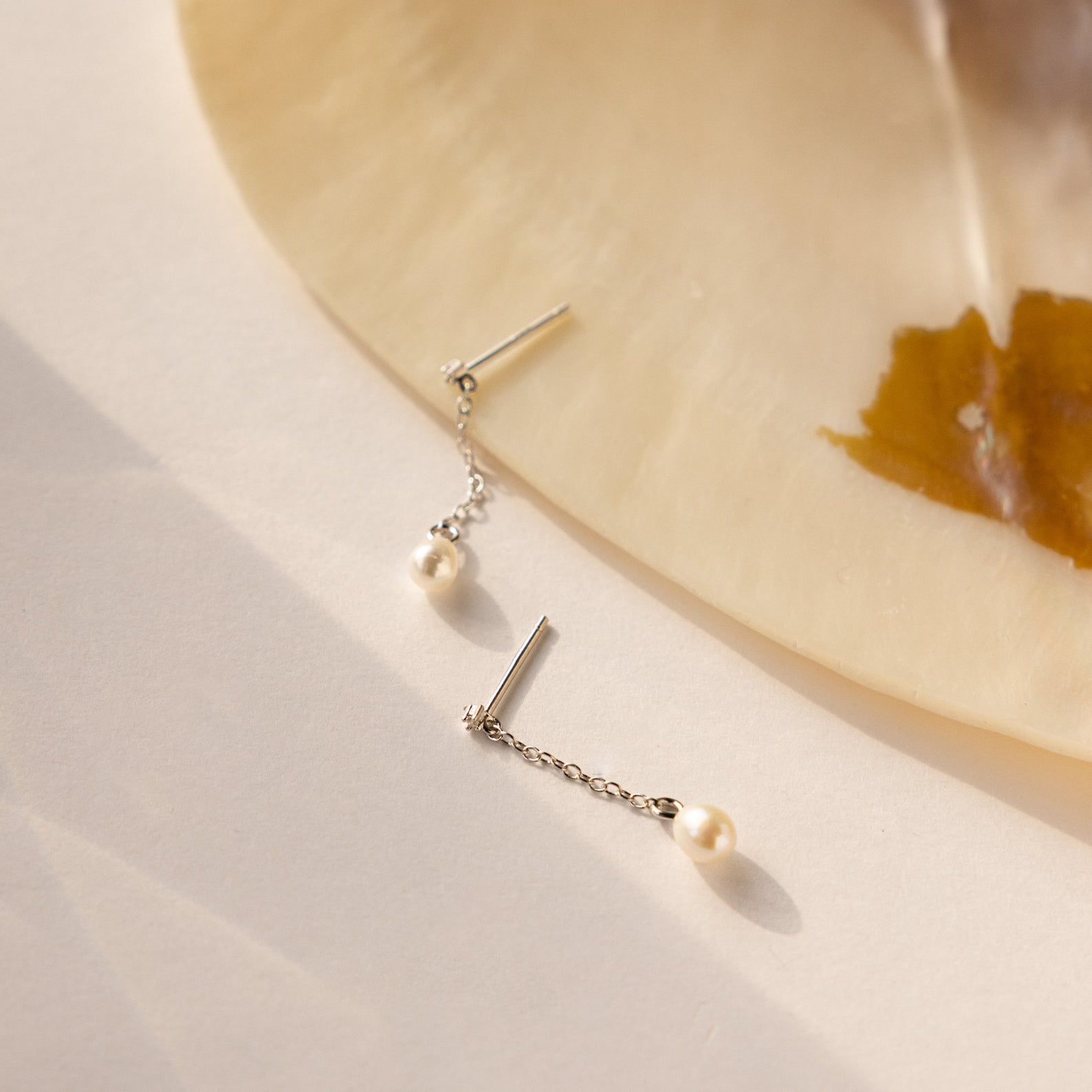The Isha Pearl Drop Earrings in Sterling Silver, shown on a light surface beside a cream and brown shell, are an ideal choice for lovers of dainty, elegant earrings.