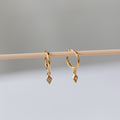 Ericka Diamond Huggies, featuring diamond-shaped hoops and dangling gemstone charms, hang delicately on a wooden stick.