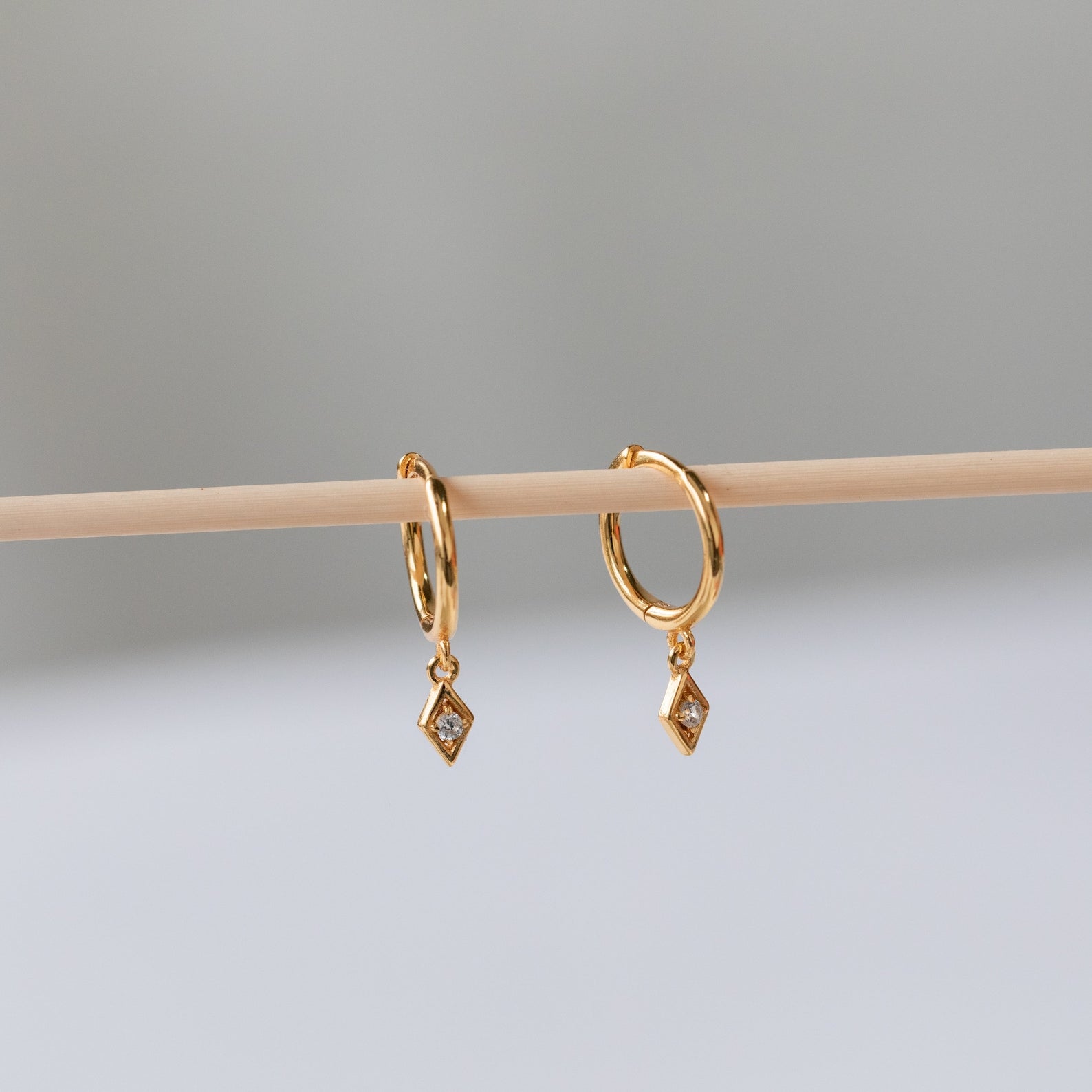 Ericka Diamond Huggies, featuring diamond-shaped hoops and dangling gemstone charms, hang delicately on a wooden stick.