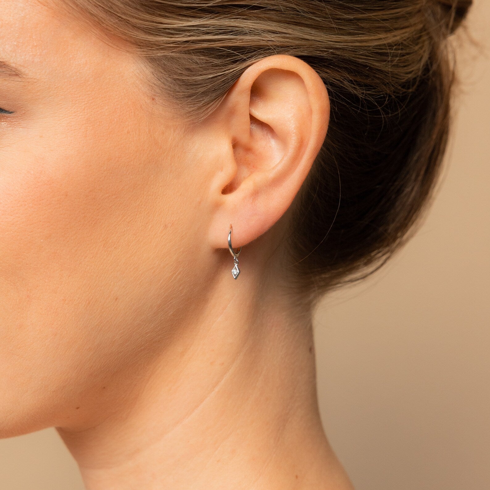 Close-up of a woman's ear wearing the Ericka Diamond Huggies in Sterling Silver, featuring sleek hoops with a delicate diamond-shaped dangling charm.