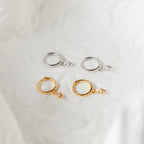 The Ericka Diamond Huggies in Sterling Silver are two pairs of huggie hoops—one gold and one silver—with diamond-shaped charms, elegantly displayed on soft white fabric.