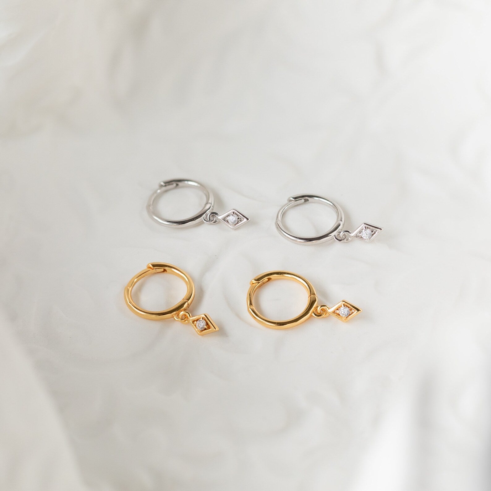 The Ericka Diamond Huggies in Sterling Silver are two pairs of huggie hoops—one gold and one silver—with diamond-shaped charms, elegantly displayed on soft white fabric.