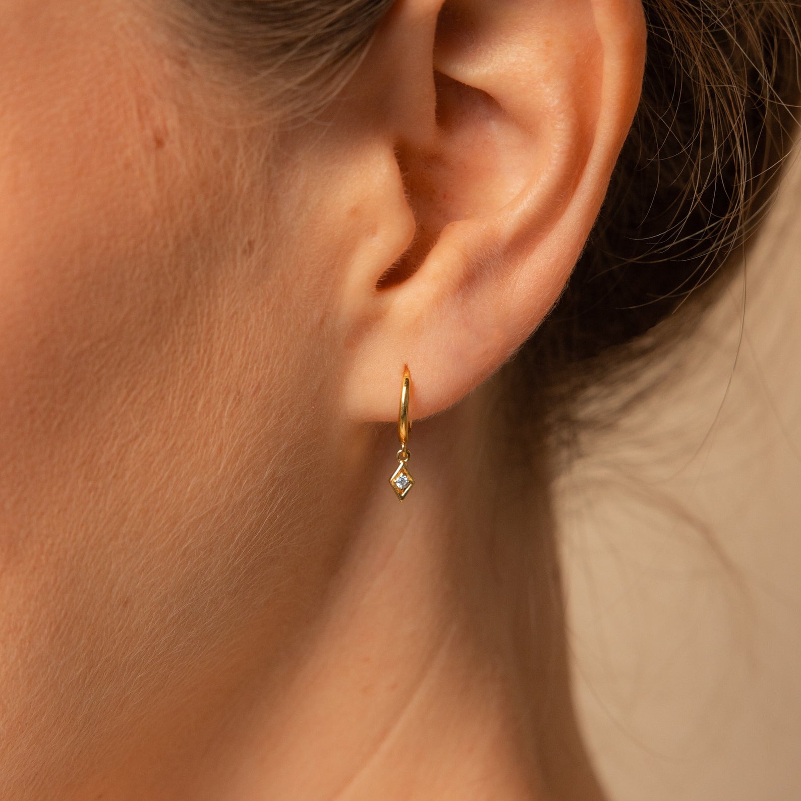 Close-up of a woman's ear wearing Ericka Diamond Huggies, featuring a dangling earring charm with a diamond-shaped pendant.