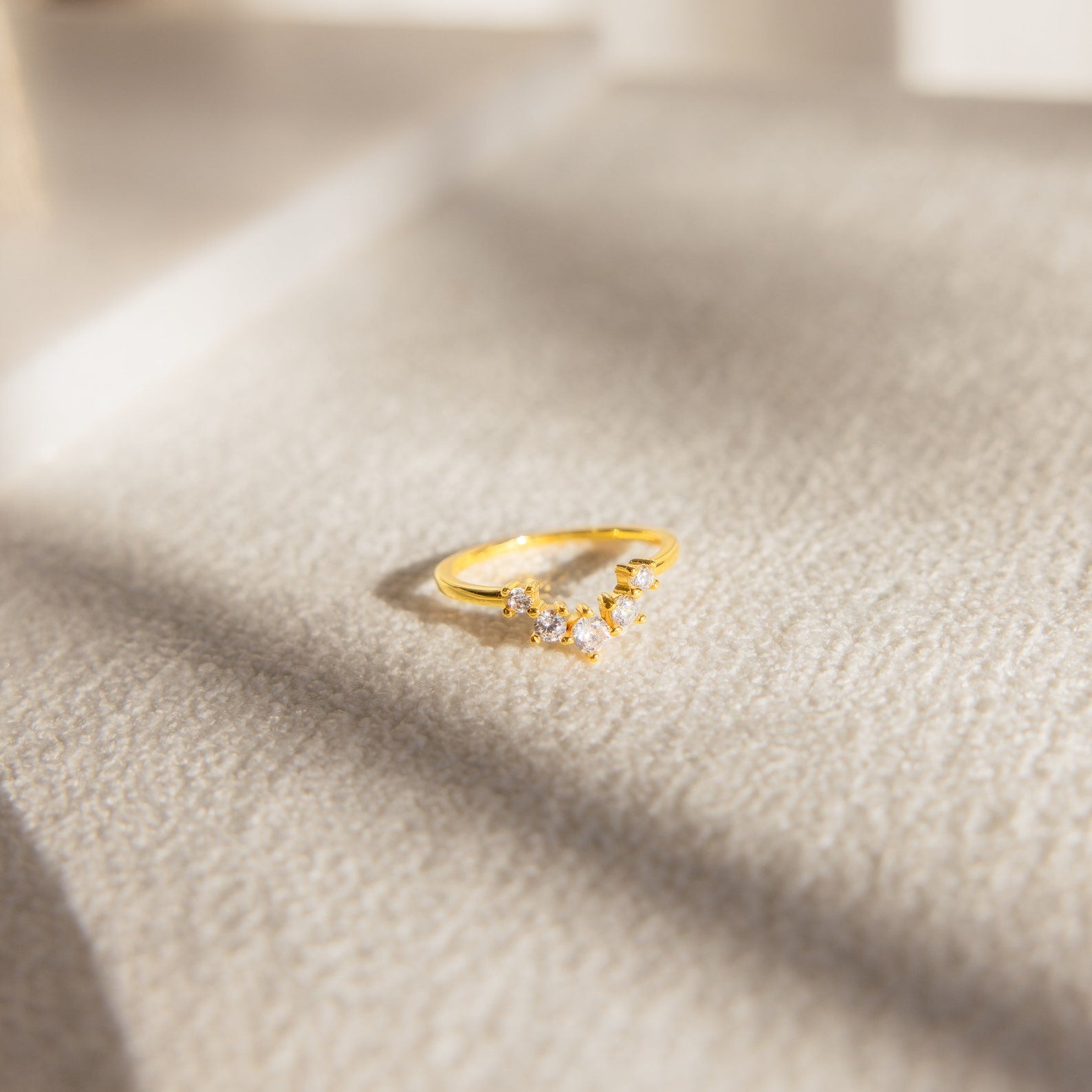 The Marina Chevron Ring, a gold engagement ring adorned with small diamonds, rests on a textured beige surface in soft sunlight.