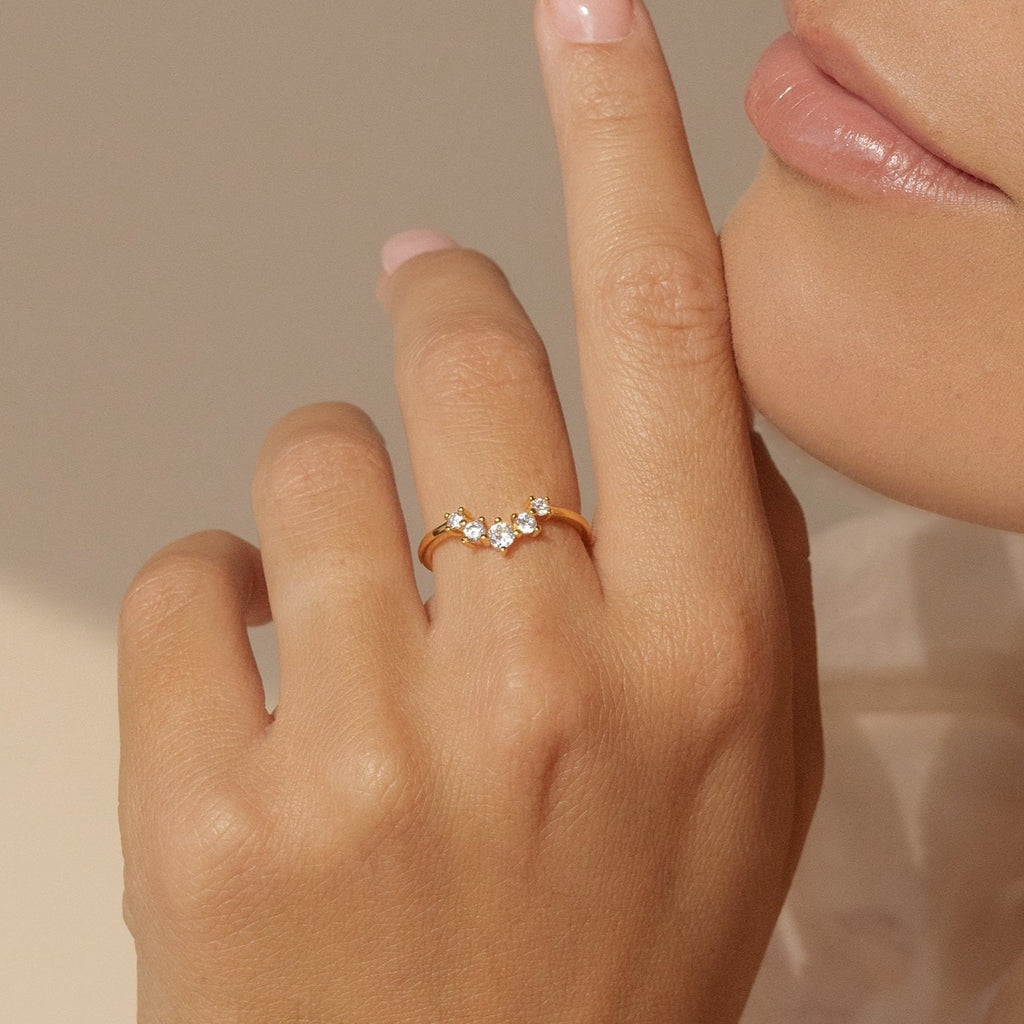 A hand adorned with the Marina Chevron Ring, showcasing three small diamonds on a delicate gold band, held near the lips against a soft neutral background.