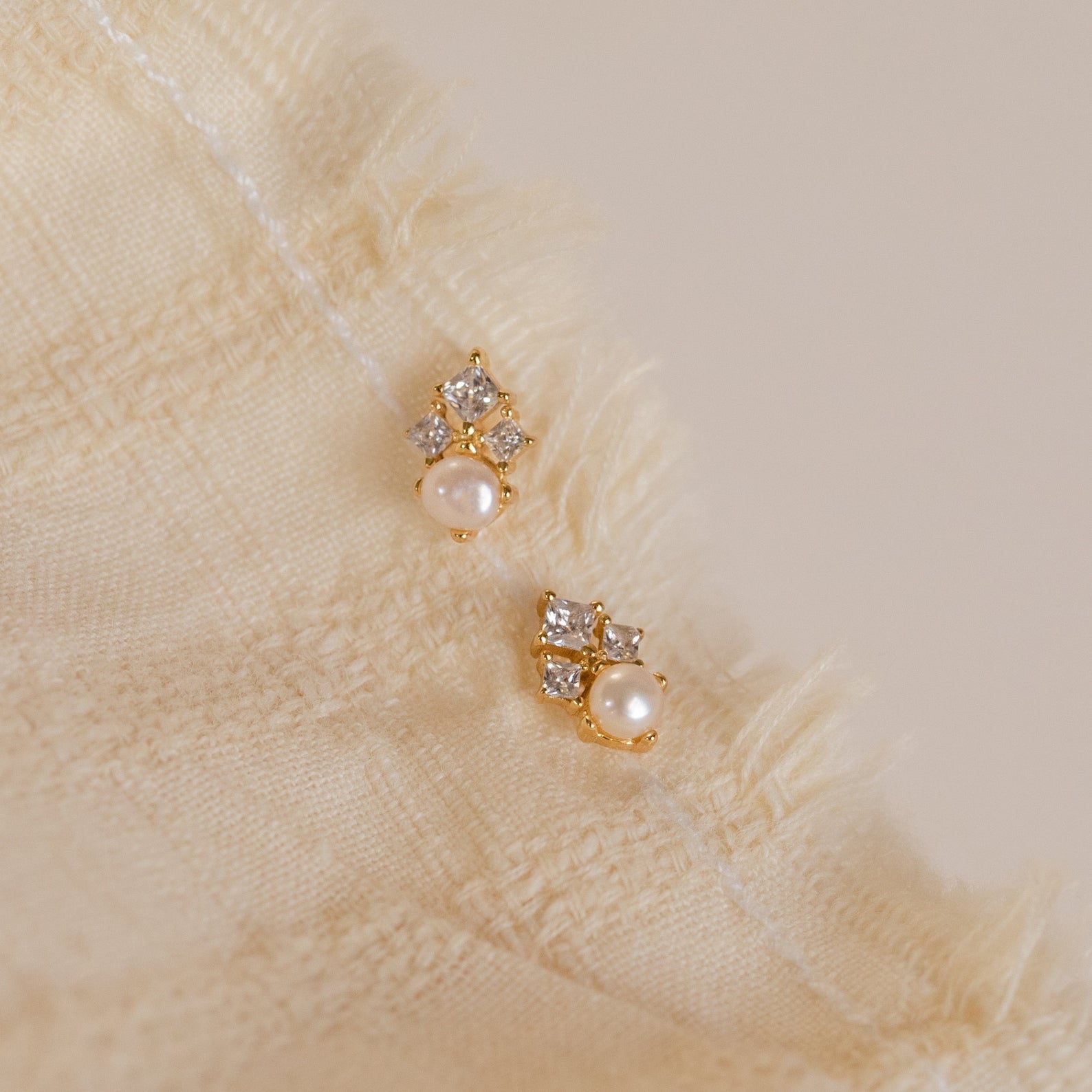 The Angelina Pearl Studs, featuring elegant pearls with gold and diamonds and a secure screw back design, are displayed on a textured cream-colored fabric background.