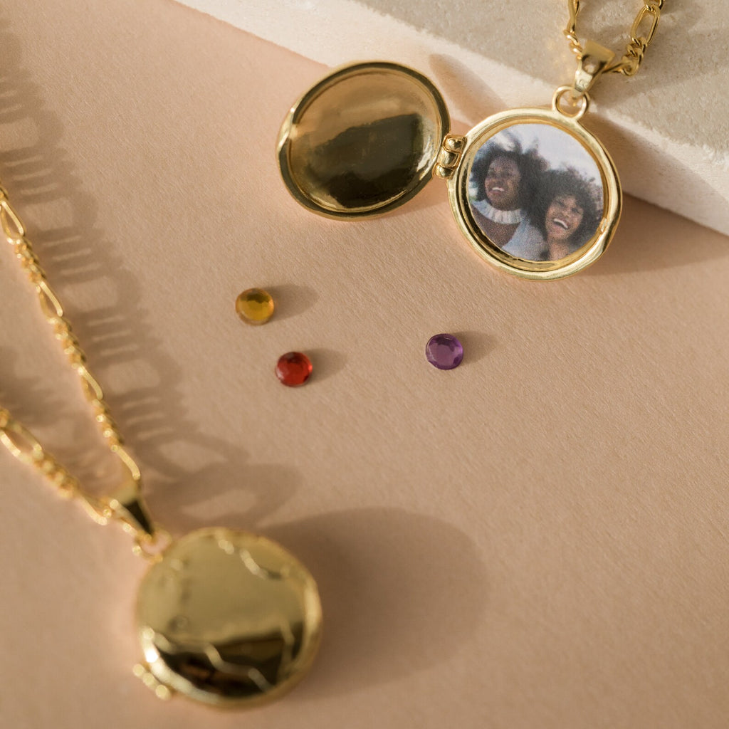 The Earth Locket Necklace rests on a beige surface, one open to reveal a photo of two smiling people with three gems nearby—creating the perfect nature-inspired keepsake to cherish your memories wherever you go.