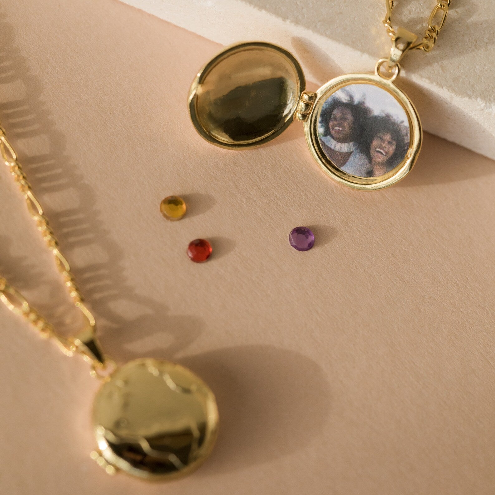 The Earth Locket Necklace rests on a beige surface, one open to reveal a photo of two smiling people with three gems nearby—creating the perfect nature-inspired keepsake to cherish your memories wherever you go.