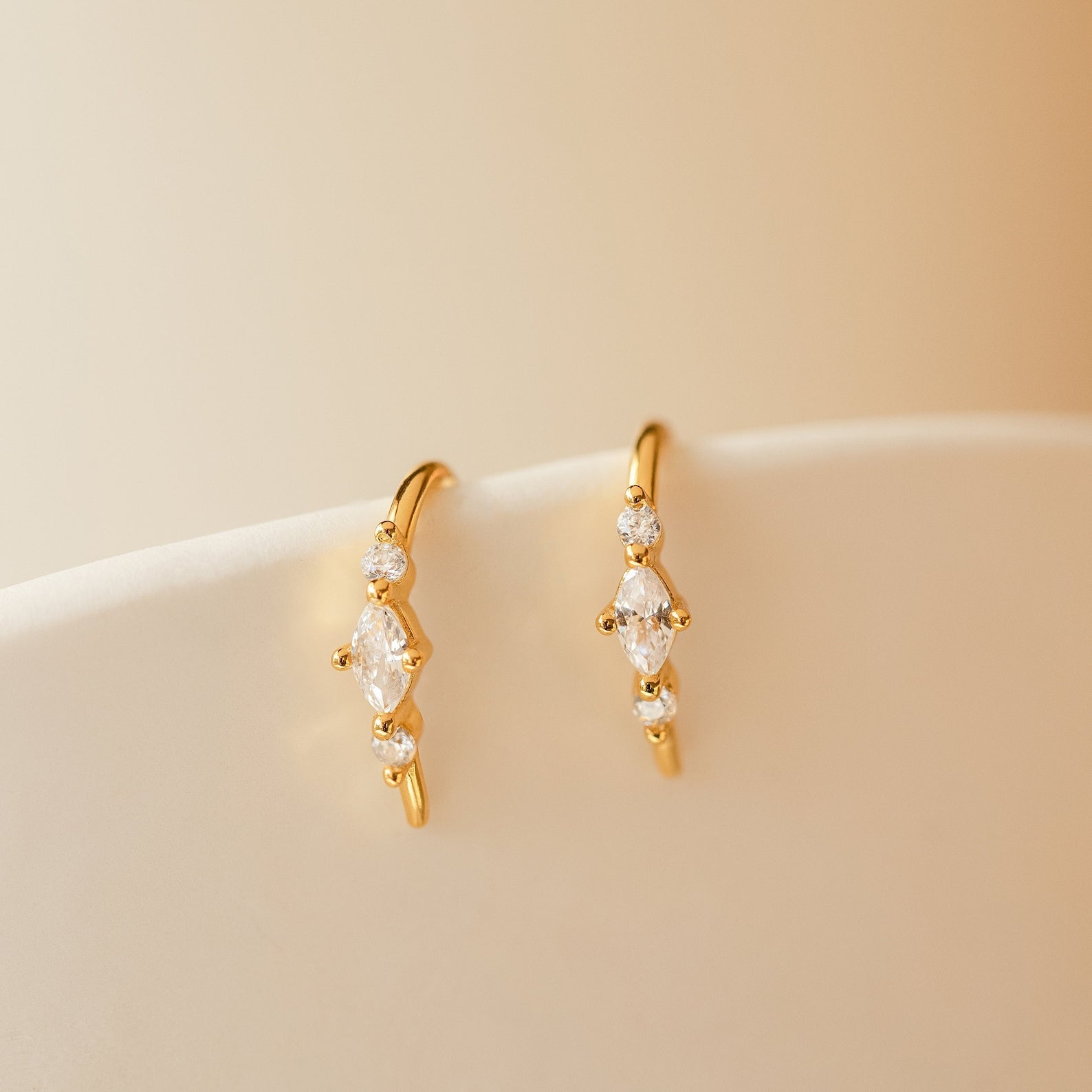 The Amaya Marquise Threaders feature gold earrings with small clear gemstones in a dainty diamond cluster, displayed on a white curved surface against a beige background—perfect for elegant jewelry lovers.