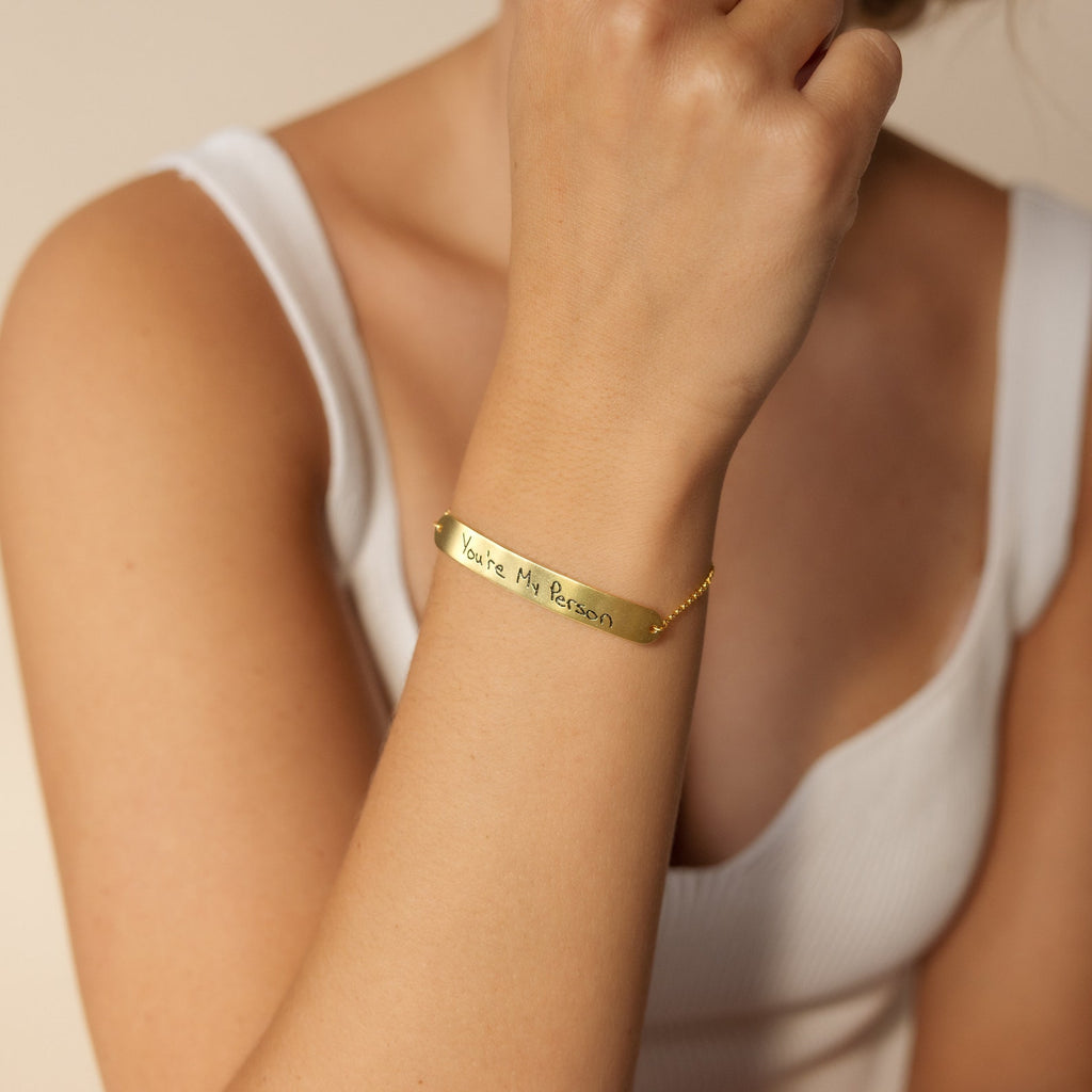 A woman in a white tank top wears a dainty Slider Handwriting Bracelet—a gold slider bar bracelet engraved with “You’re My Person.”.