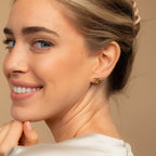 A woman smiles, showing off gold Heart Knot Studs with a delicate knot design. Her hair is styled back, and she wears a light-colored top.