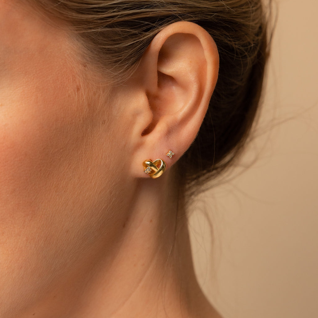 Close-up of a woman's ear with two earrings: a diamond charm stud and the Heart Knot Studs, displaying their intricate gold knot design.