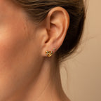 Close-up of a woman's ear with two earrings: a diamond charm stud and the Heart Knot Studs, displaying their intricate gold knot design.