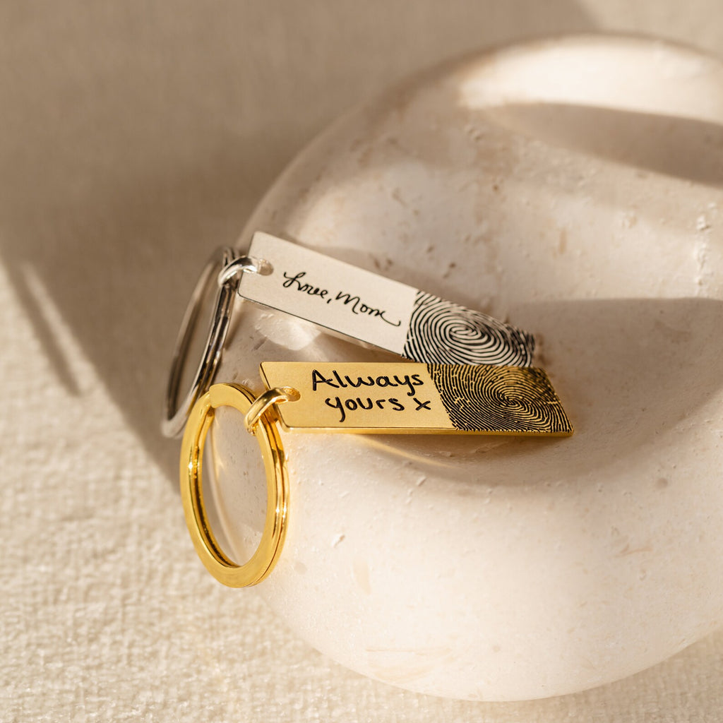 The Fingerprint Bar Keychain features two engraved keychains—one silver with "Love, Mom," and one gold with "Always yours x"—both with your actual fingerprints. This personalized keepsake is a touching reminder of your unique bond.