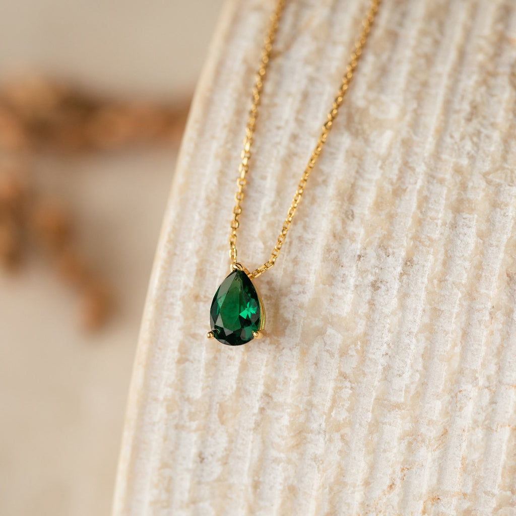 The Ophelia Birthstone Necklace features a teardrop-shaped green stone pendant on a gold chain, elegantly displayed on a textured beige surface for a minimalist look.