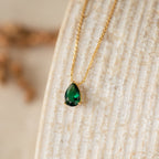 The Ophelia Birthstone Necklace features a teardrop-shaped green stone pendant on a gold chain, elegantly displayed on a textured beige surface for a minimalist look.