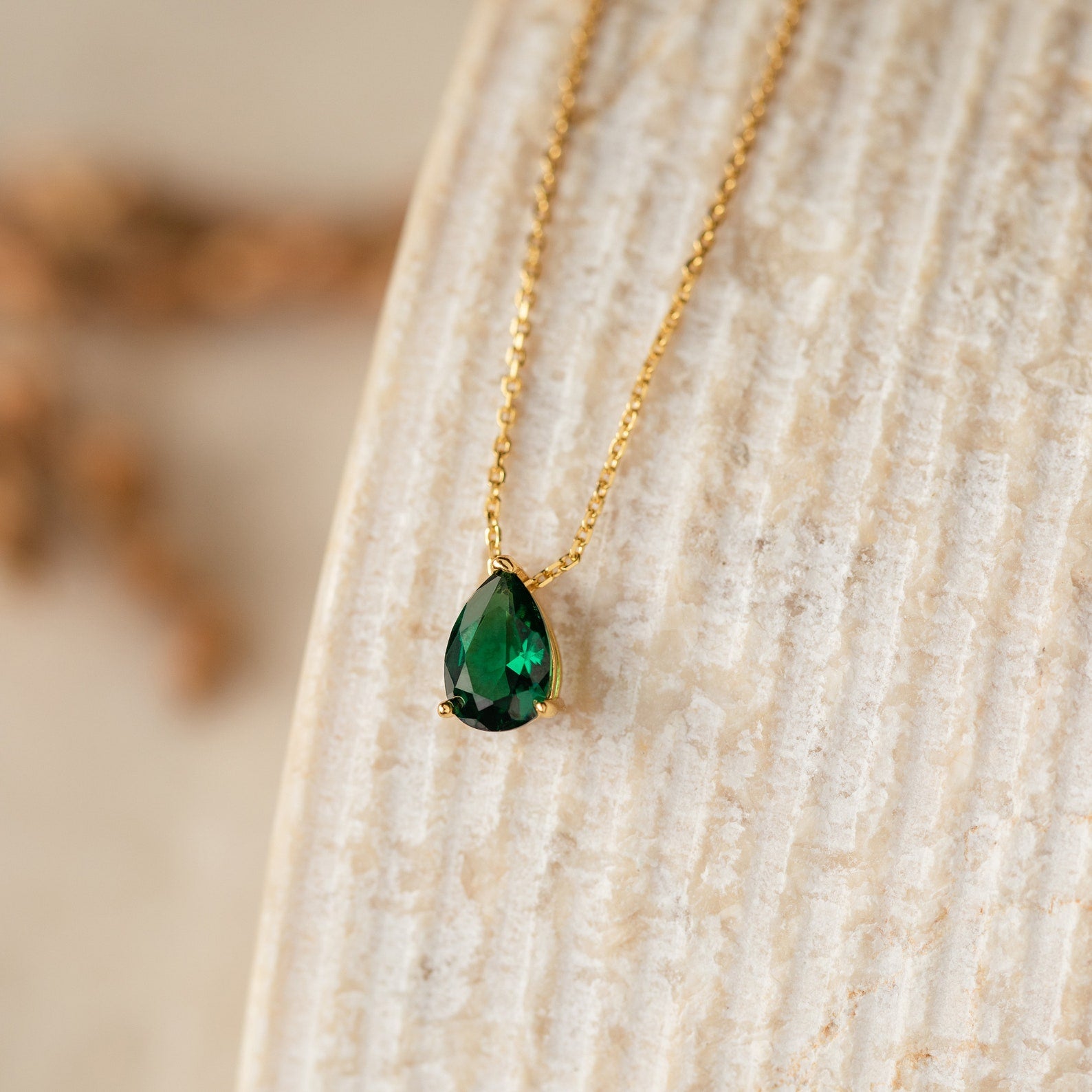 The Ophelia Birthstone Necklace features a teardrop-shaped green stone pendant on a gold chain, elegantly displayed on a textured beige surface for a minimalist look.