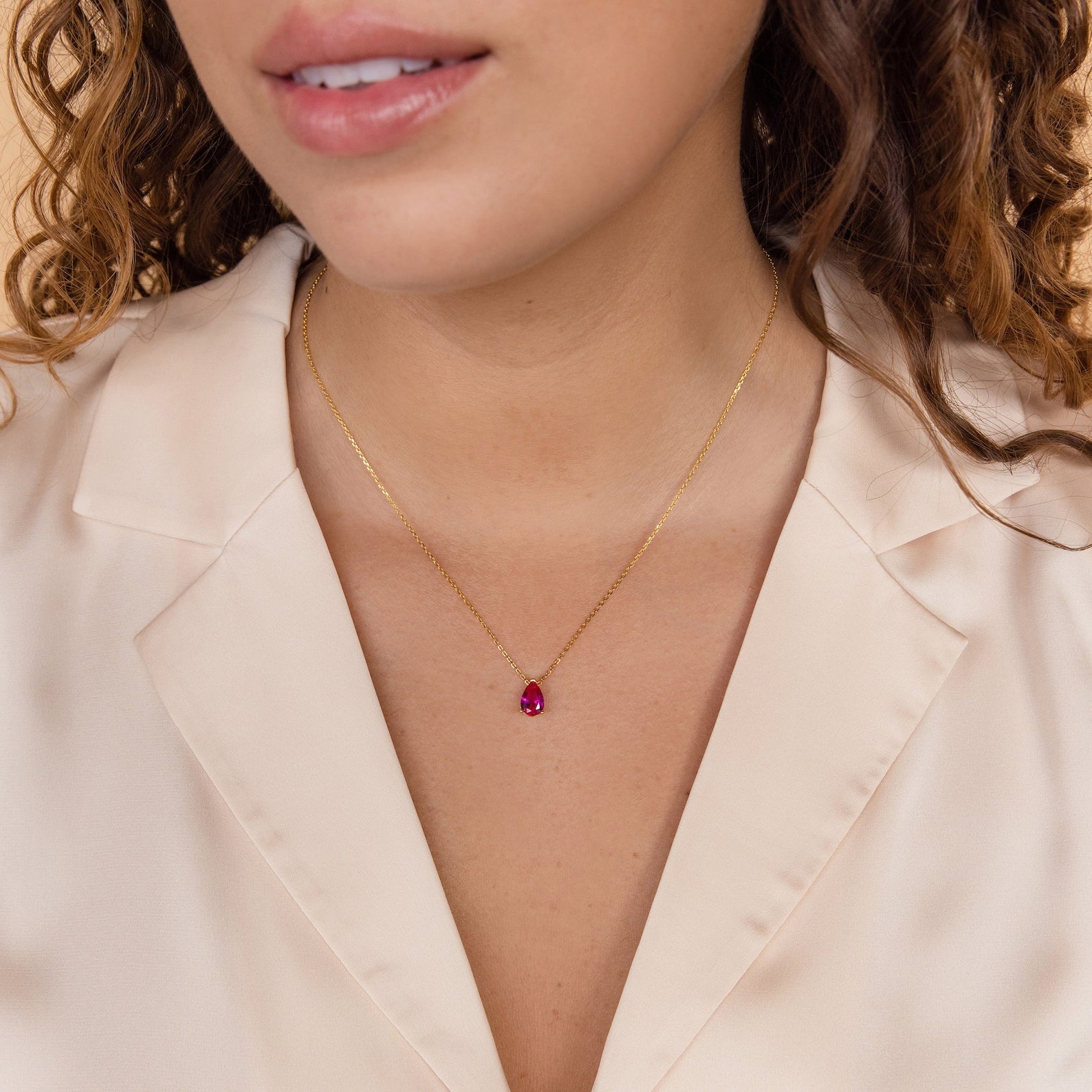 A woman in a cream-colored blouse wears the Ophelia Birthstone Necklace—a minimalist gold chain featuring a small red birthstone pendant.