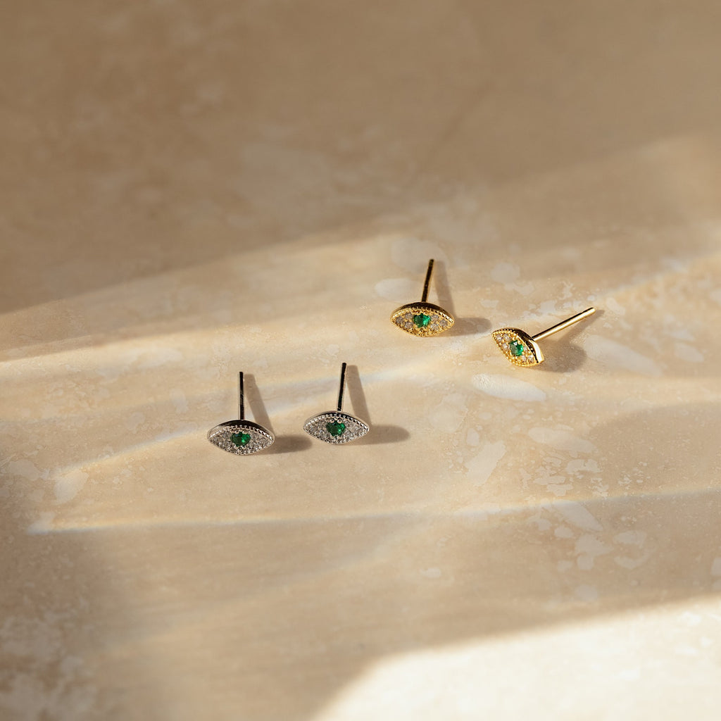 The Haven Emerald Eye Studs, a set of four elegant earrings with emerald stones, are showcased on a beige textured surface in gentle sunlight.
