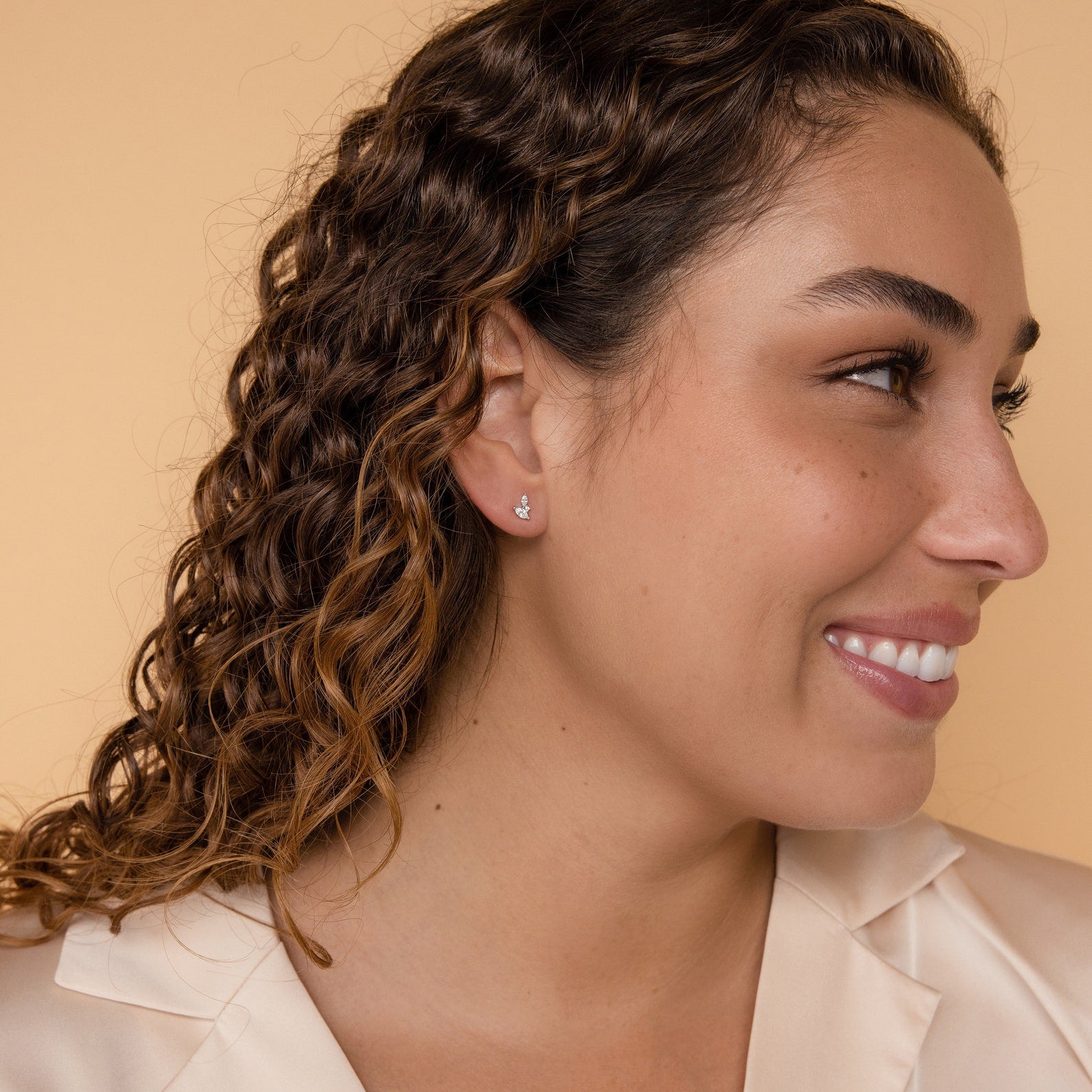 Woman with curly hair smiles against a peach background, wearing a cream blazer and Flora Marquise Studs in Sterling Silver.