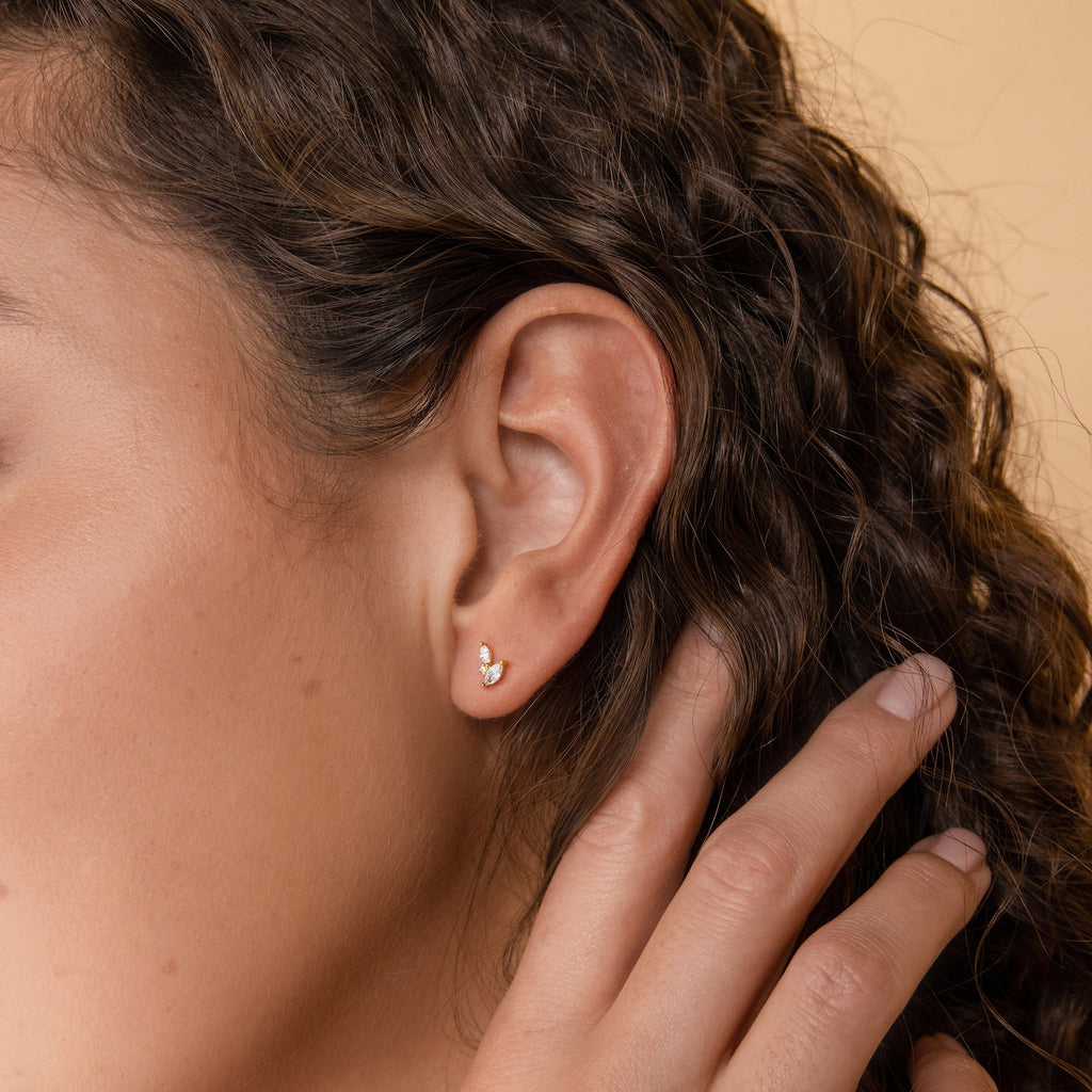 Close-up of a person with curly brown hair wearing Flora Marquise Studs in 18K Gold and gently touching their ear.