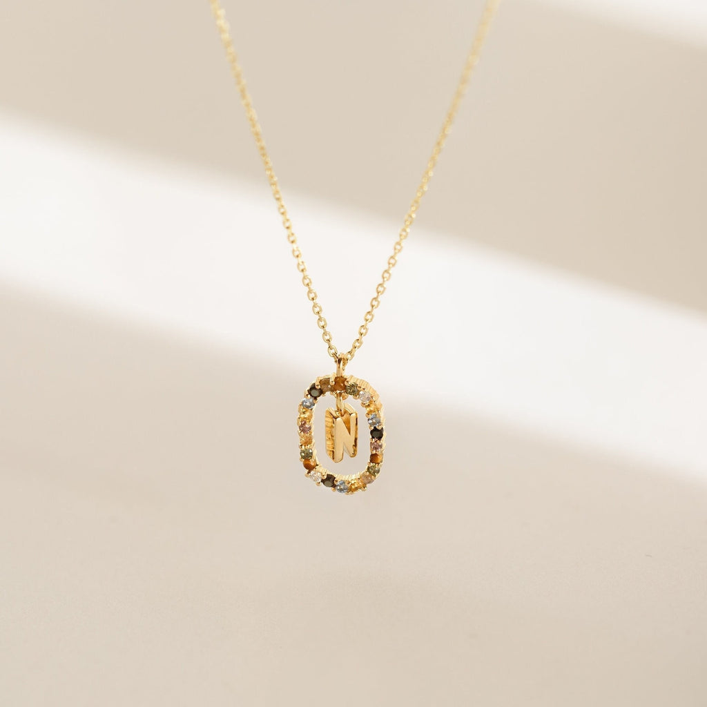 The Rainbow Initial Necklace features a gold chain with an oval pendant showcasing the letter M, accented by small multicolored stones—an eye-catching accessory for a personalized touch.