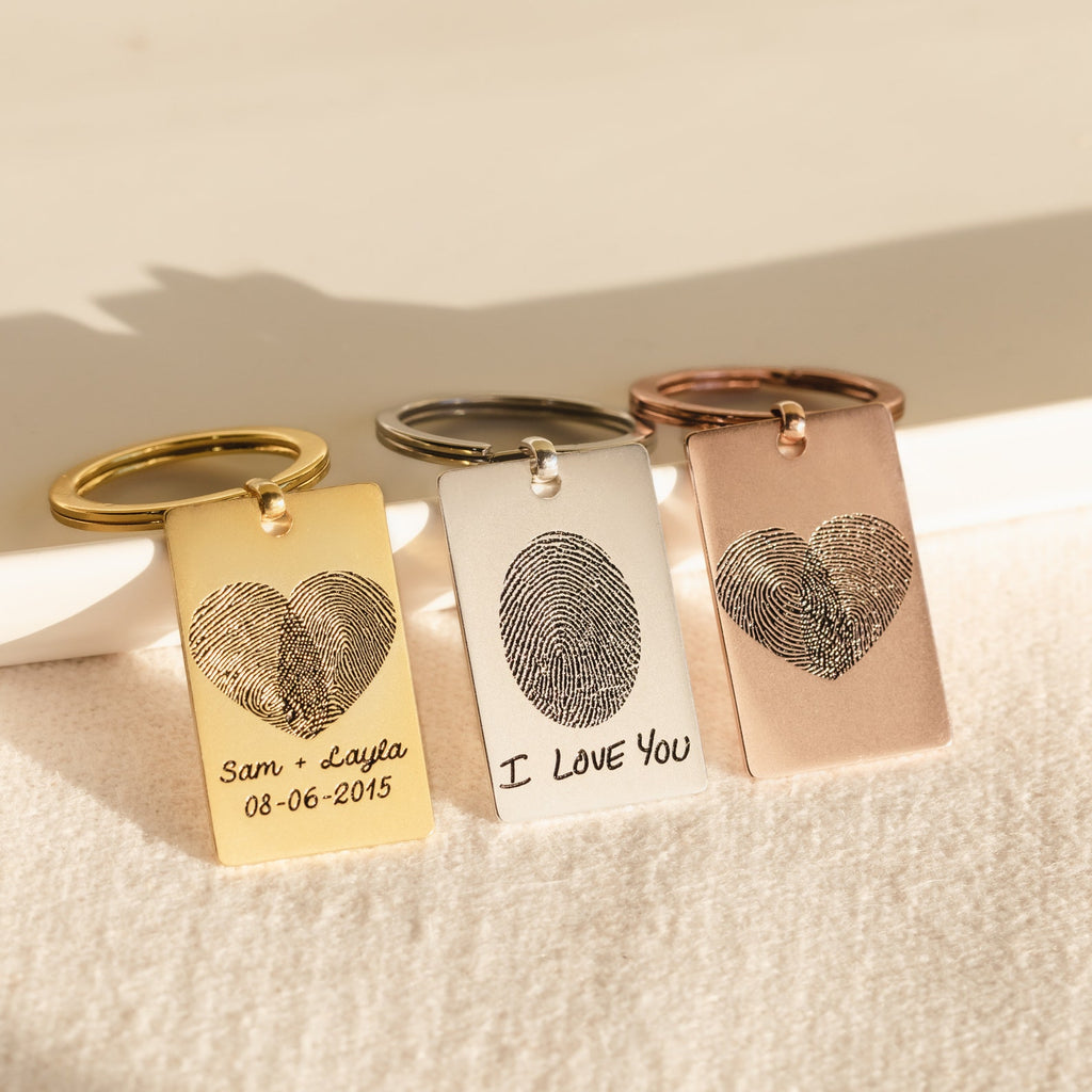 Three engraved Fingerprint Rectangle Keychains with heart motifs and custom messages rest on a light fabric, making each piece a unique personalized keepsake.