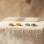 The Opal Inlay Huggies Set features two pairs of huggie hoop earrings—one gold, one silver—displayed on a textured white surface in sunlight, with opal inlays catching the light and shimmering beautifully.