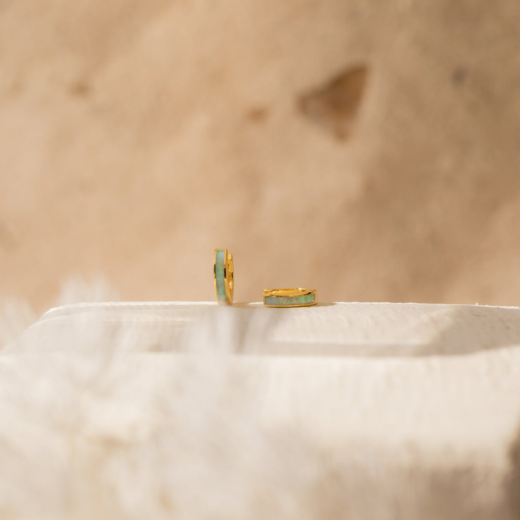 The Opal Inlay Huggies Set features two gold hoops with green opal accents, displayed on a light textured surface with a beige blurred background, highlighting their shimmering opal hues.