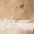 The Opal Inlay Huggies Set features two gold hoops with green opal accents, displayed on a light textured surface with a beige blurred background, highlighting their shimmering opal hues.