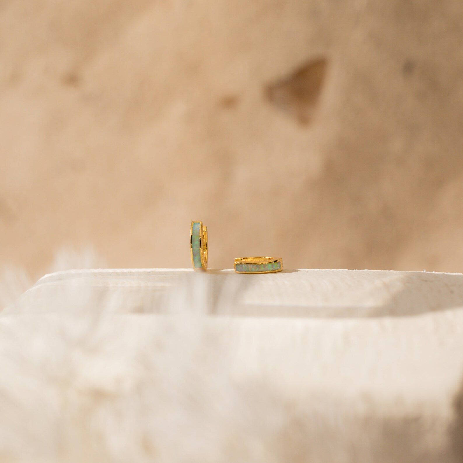 The Opal Inlay Huggies Set features two gold hoops with green opal accents, displayed on a light textured surface with a beige blurred background, highlighting their shimmering opal hues.