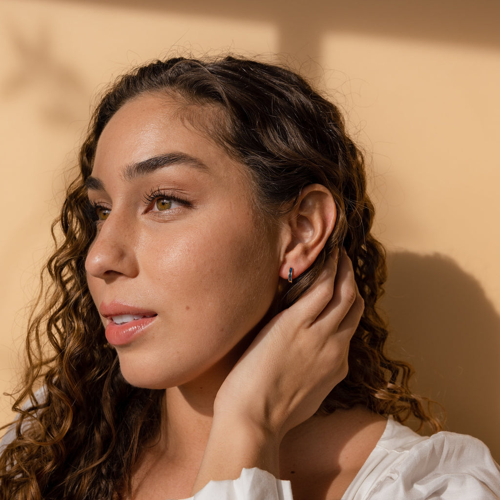 A woman with curly hair in a white top touches her ear, showcasing the Opal Inlay Huggies Set against a beige background in soft sunlight.