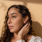 A woman with curly hair in a white top touches her ear, showcasing the Opal Inlay Huggies Set against a beige background in soft sunlight.