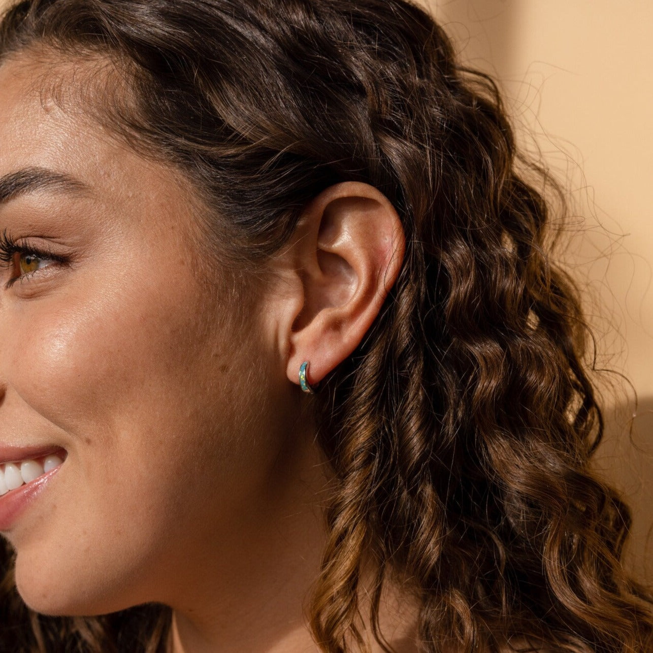 A woman with curly hair smiles brightly, her look enhanced by the Opal Inlay Huggies Set.