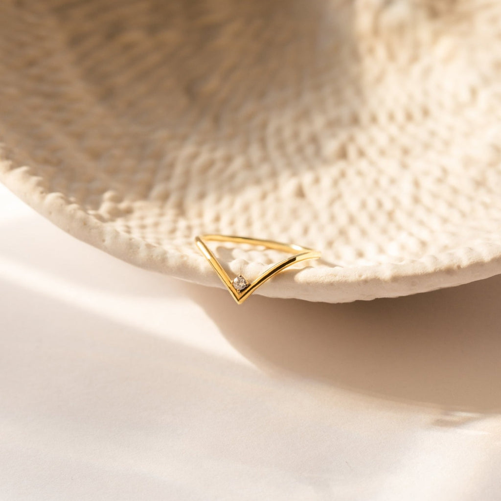 The Chevron Birthstone Ring, a gold triangular gemstone ring with a small diamond, rests on the edge of a textured ceramic dish.
