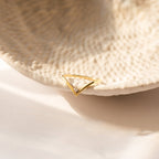 The Chevron Birthstone Ring, a gold triangular gemstone ring with a small diamond, rests on the edge of a textured ceramic dish.