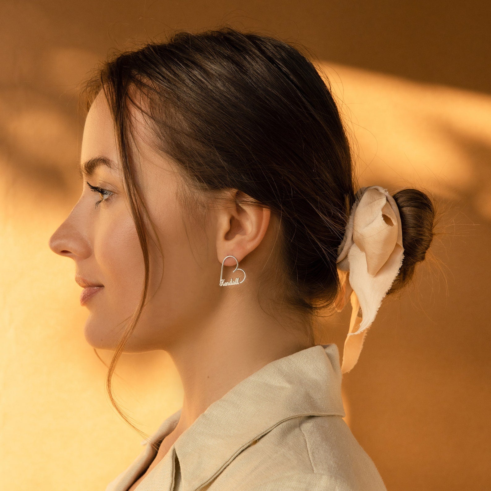 A woman in profile with her hair tied back in a silk ribbon, wearing London Heart Name Earrings—personalized heart-shaped hoops featuring elegant custom names.