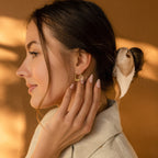 A woman with a white ribbon in her hair touches her London Heart Name Earrings, gazing thoughtfully to the side.