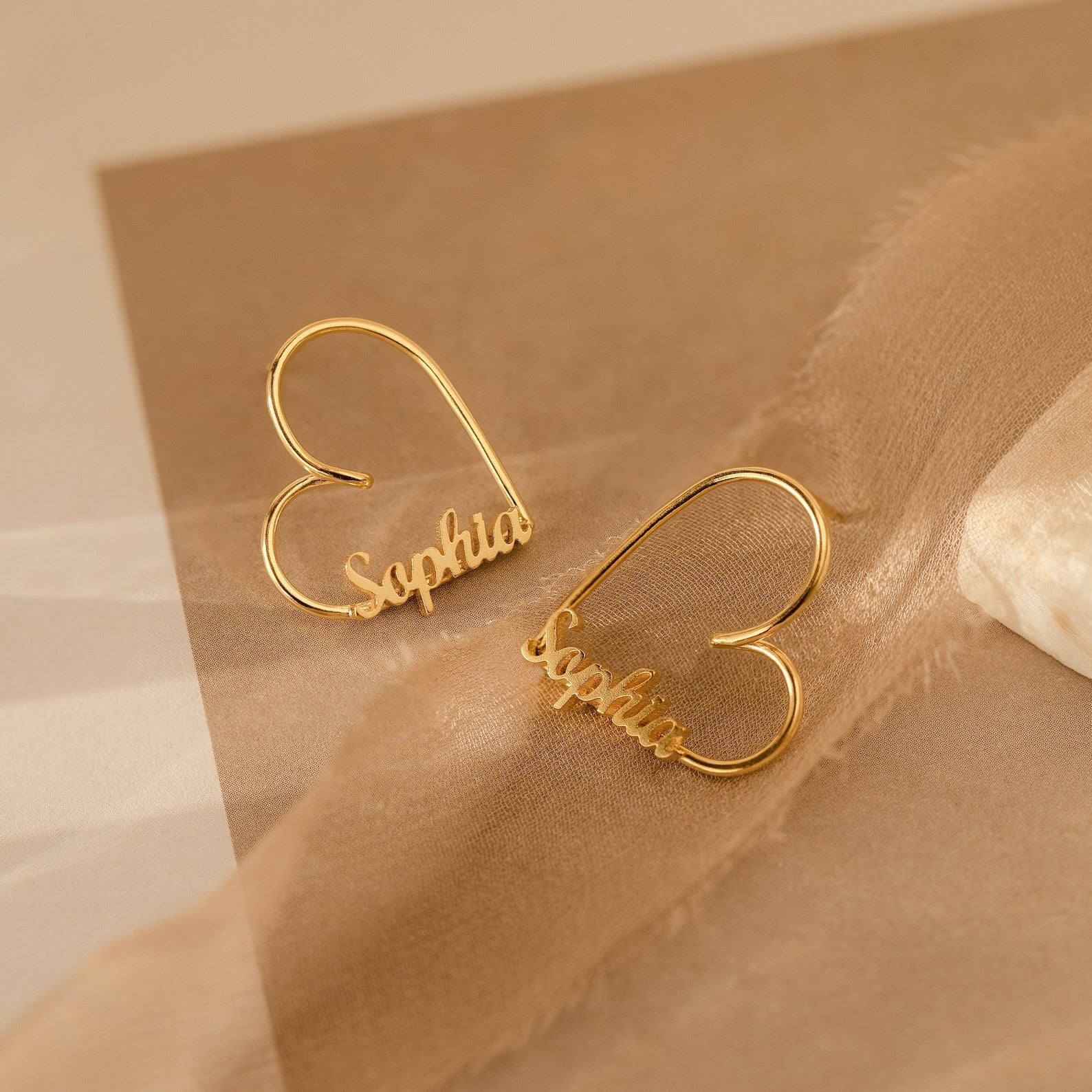 London Heart Name Earrings in gold, heart-shaped hoops featuring the name Sophia, are displayed on a beige and brown soft fabric background—making them an ideal personalized gift.