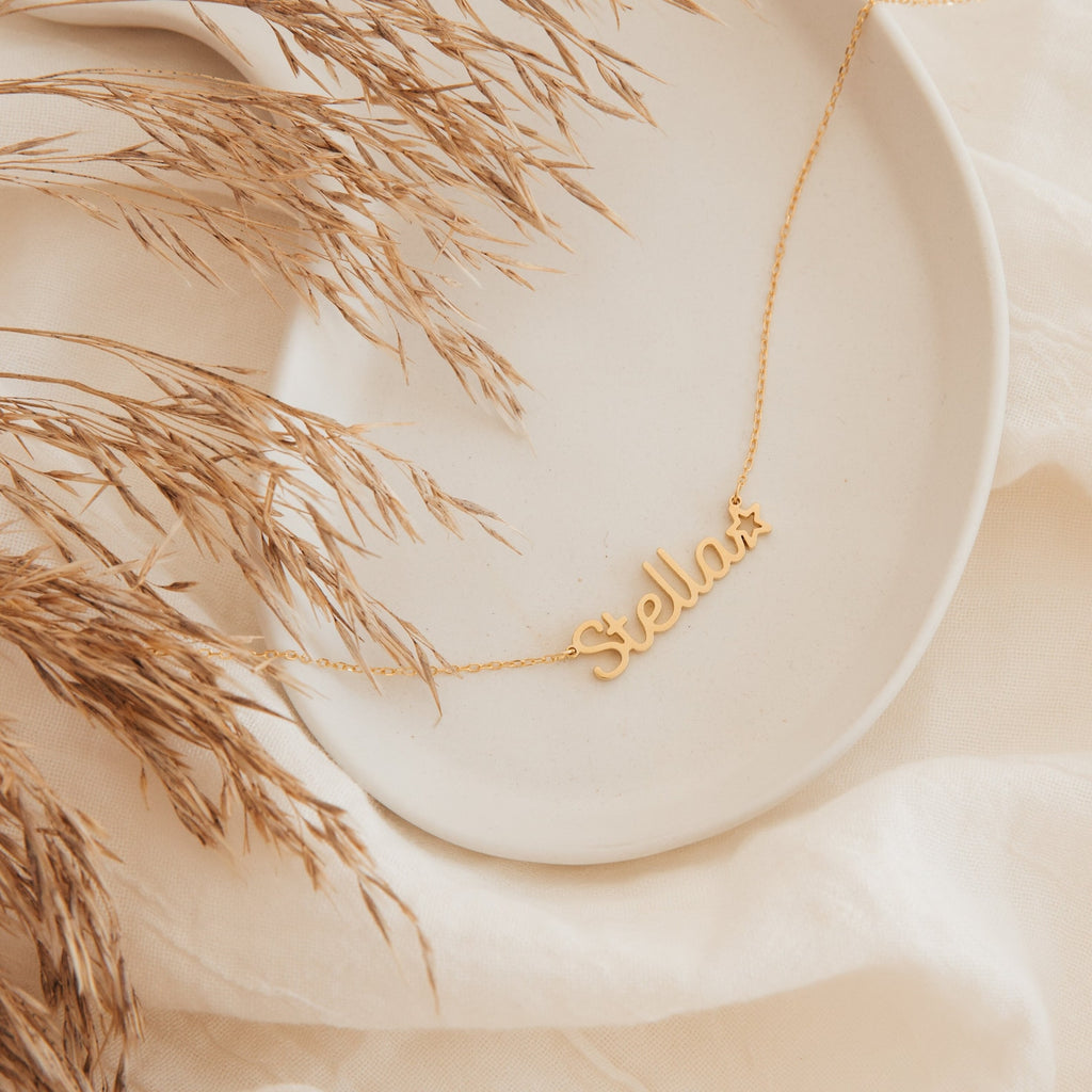 The Maine Kid's Name Necklace, featuring "Stella" and a star charm, is displayed on a cream plate with dried grass—making it a charming personalized piece and the perfect kids’ jewelry gift.