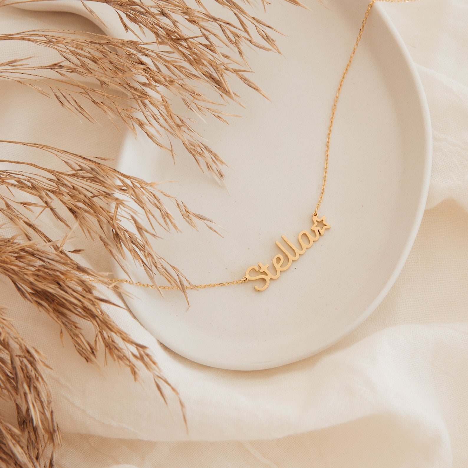 The Maine Kid's Name Necklace, featuring "Stella" and a star charm, is displayed on a cream plate with dried grass—making it a charming personalized piece and the perfect kids’ jewelry gift.