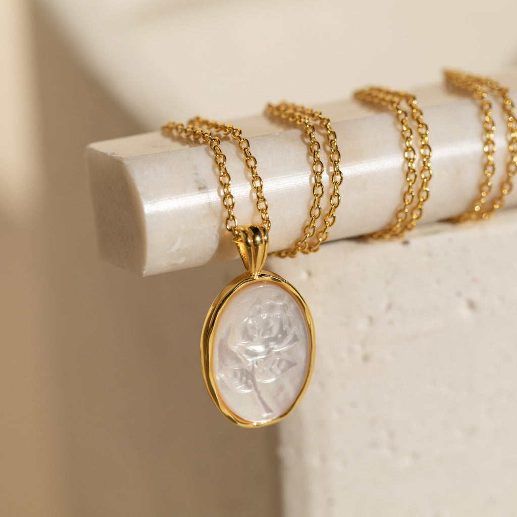 The Vintage Rose Necklace features a gold chain and an oval translucent pendant engraved with a rose, displayed on a cream surface—a lovely flower necklace perfect for Mother’s Day.