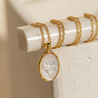 The Vintage Rose Necklace features a gold chain and an oval translucent pendant engraved with a rose, displayed on a cream surface—a lovely flower necklace perfect for Mother’s Day.